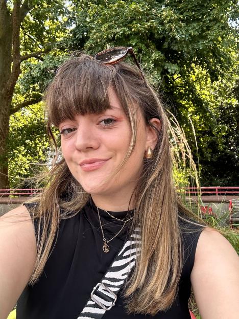 Cyann Fielding smiling at the camera outdoors, wearing a black sleeveless top, gold earrings, and layered gold necklaces, with sunglasses perched on her head.