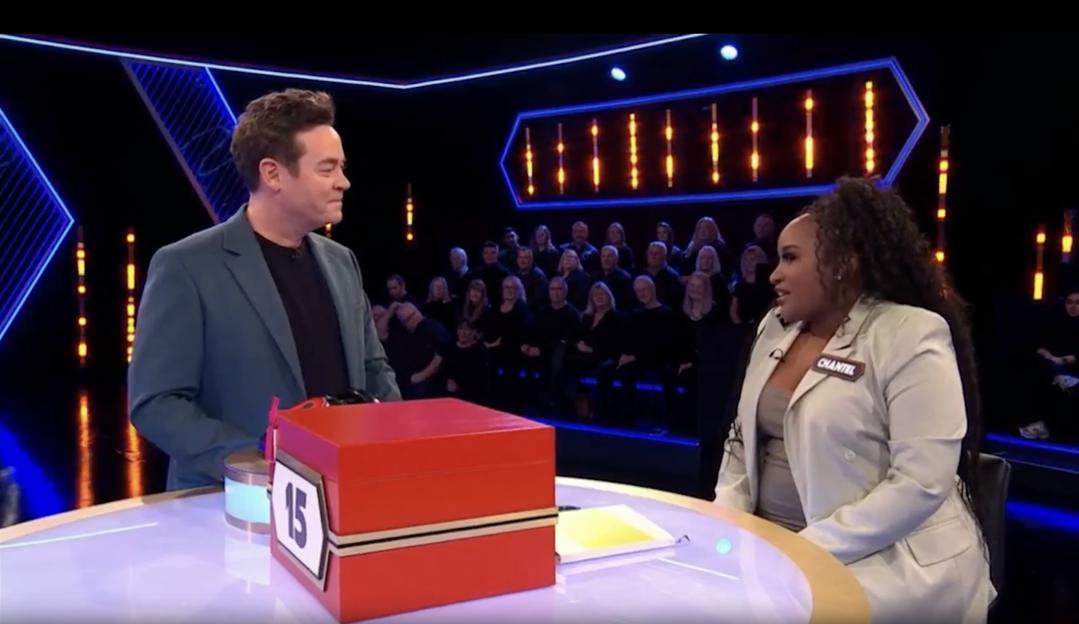 A TV game show host and a female contestant sit opposite each other at a brightly lit table with a red box labeled "15" between them.