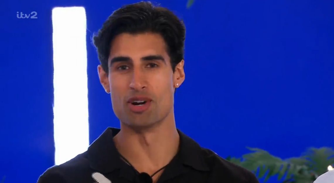 A man with dark hair and a black shirt talking, with an ITV2 logo in the background.