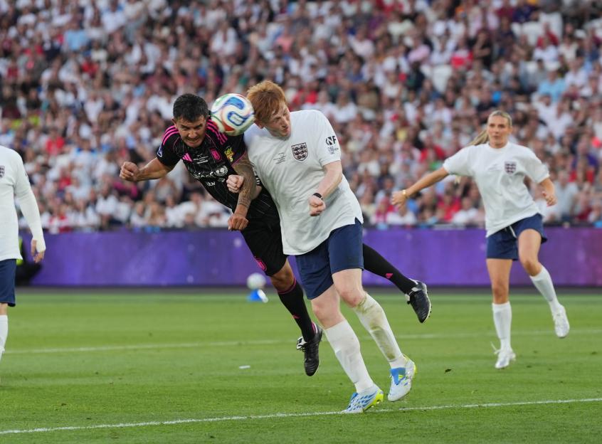 Two male soccer players head a ball while a female player runs in the background at the London Stadium during Soccer Aid for UNICEF 2026.