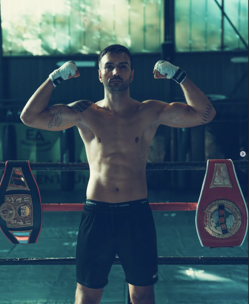 A shirtless boxer with white hand wraps poses in a boxing ring with two championship belts on either side of him.
