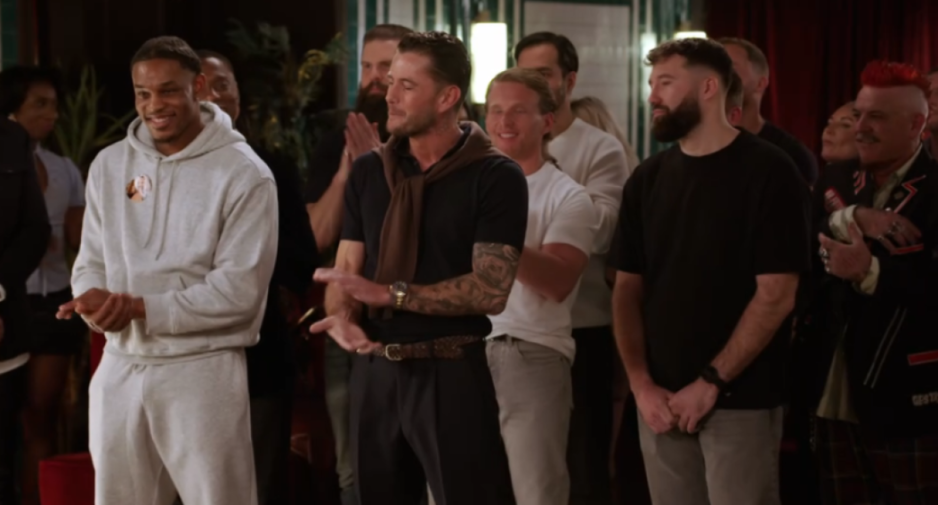 A group of men stand and clap in a dimly lit room, with one man in the foreground wearing a grey sweatsuit and another man with tattoos on his arms, a brown sweater over his shoulders, and dark pants.