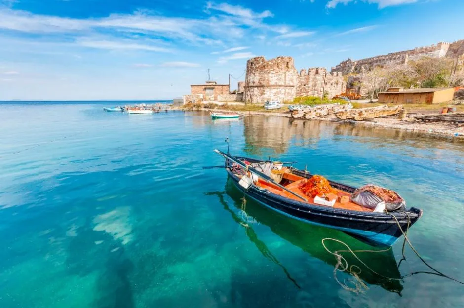 A small fishing boat moored in the calm, blue waters of the old harbour in Mytilini, Lesvos Island, with a medieval fortress in the background.