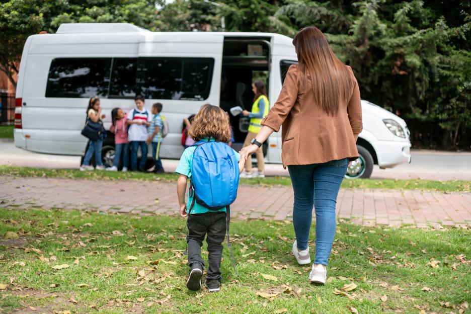A mother walks her son to the school bus, holding his hand.