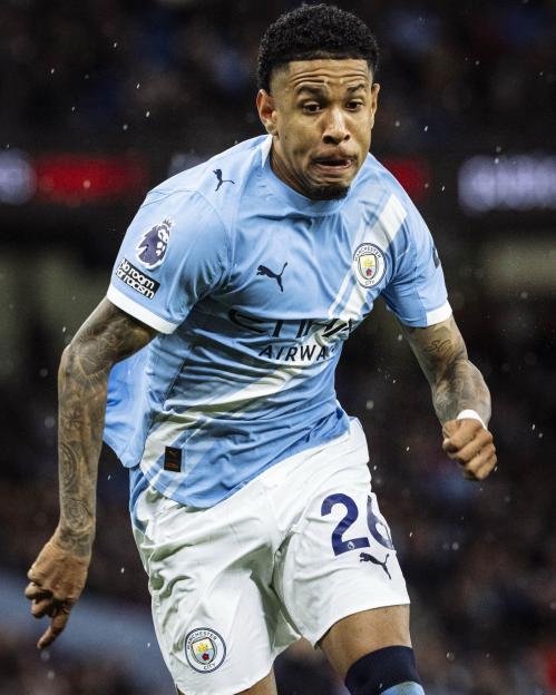 A man wearing a light blue Manchester City soccer jersey and white shorts with number 26 on them runs across a field.