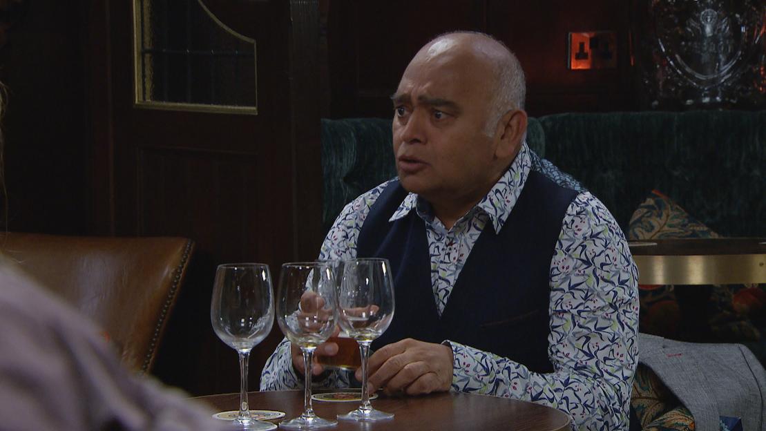 Rishi Sharma sitting at a table with three empty wine glasses and a drink in his hand, looking concerned.