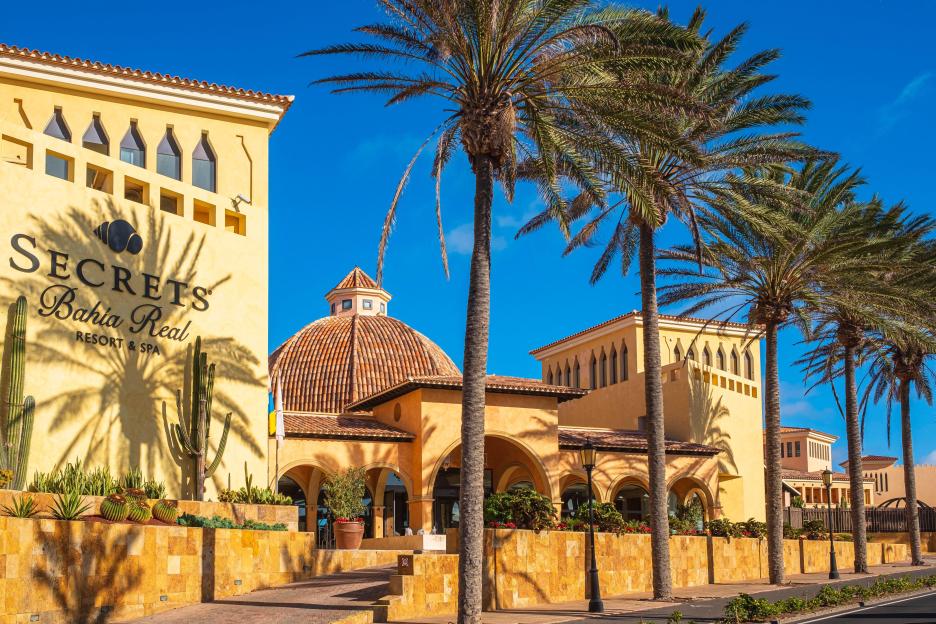 The exterior of Secrets Bahia Real Resort & Spa in Corralejo, Fuerteventura, Spain, featuring yellow buildings, a tiled dome, and palm trees.