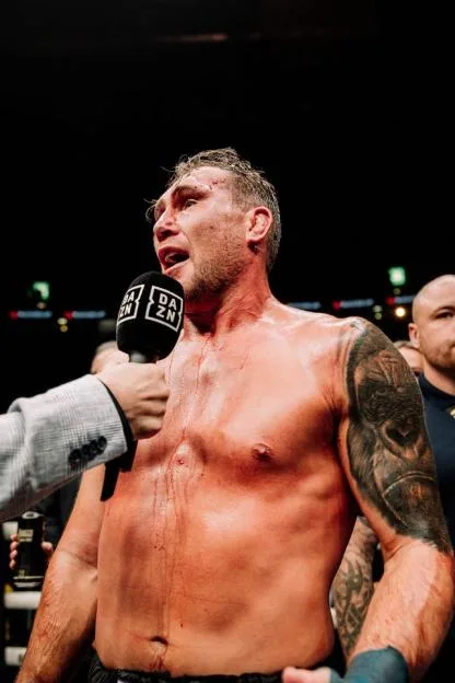 A male boxer, shirtless and covered in blood, giving an interview after a fight.