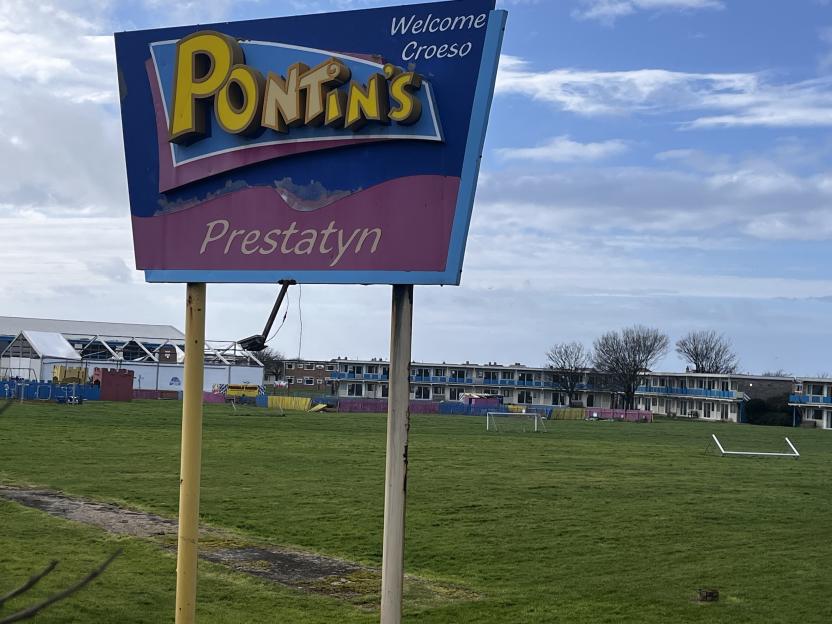 Sign for the former Pontins holiday park in Prestatyn, showing the park's name, "Pontins Prestatyn," over a grassy field with buildings in the background.
