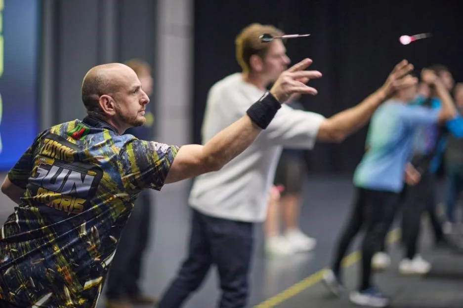 A darts player in a patterned shirt with "Tommie 'The Gun' Morris" on the back throws a dart.