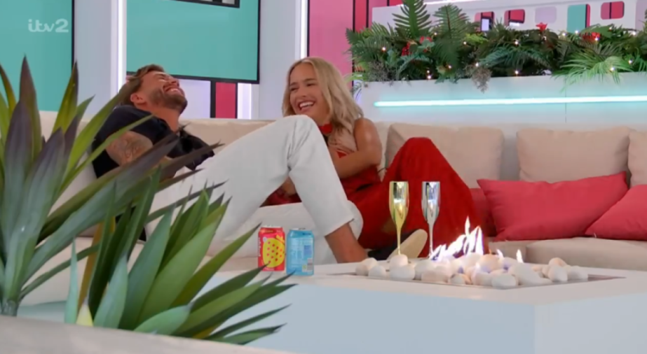Two people sitting on a white sofa and laughing, with a fireplace, plants, and drinks in the foreground.