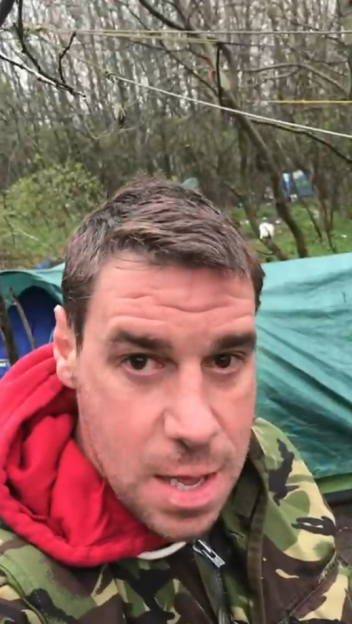 A man in a camouflage jacket and red hoodie stands outdoors with tents and trees in the background.
