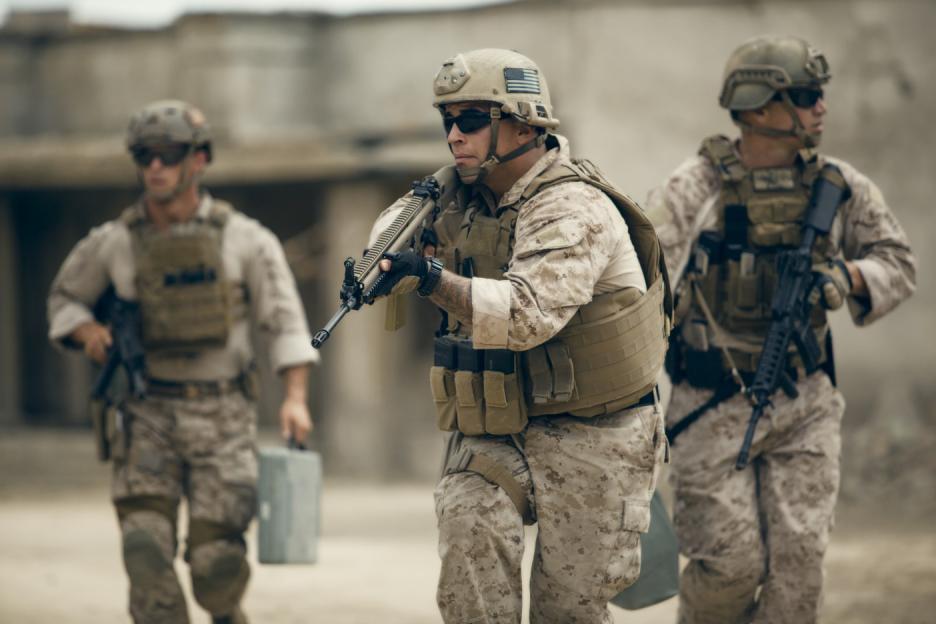 Three soldiers in desert camouflage and tactical gear walking in a blurred, dusty outdoor environment.