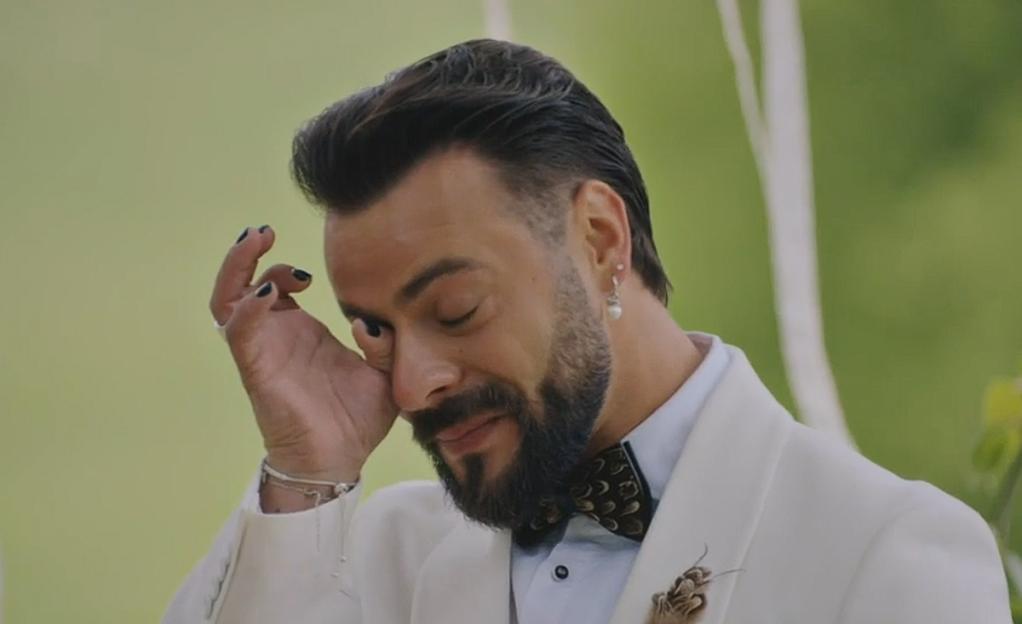 Man in a white suit with black fingernails wiping his eye while crying.