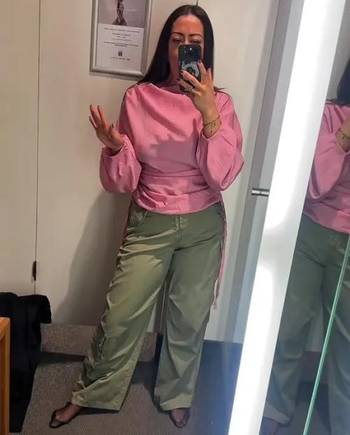 A woman in a fitting room trying on a pink top and olive green cargo pants.