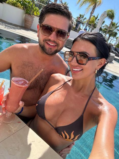 A man and a woman in sunglasses in a swimming pool, with the woman holding a pink drink.