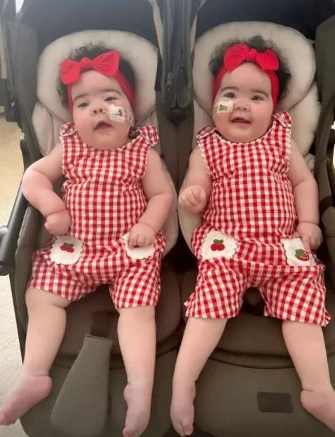 Twin baby girls in matching red and white outfits sitting side by side in a double stroller.