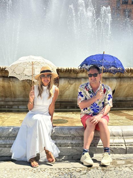 Amanda Holden and Alan Carr sitting by a fountain, holding parasols.
