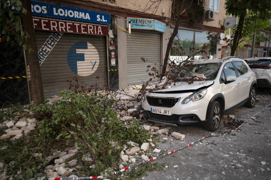 A white SUV parked in front of closed storefronts, damaged with a shattered windshield, debris, and fallen tree branches on and around it.