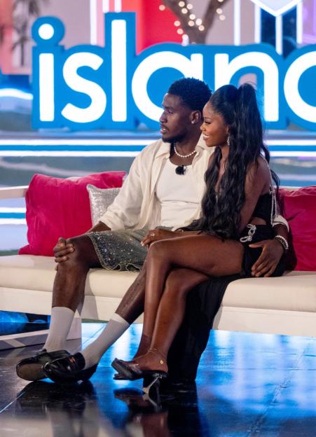 A man and a woman sit together on a white couch with a glowing "Island" sign in the background.