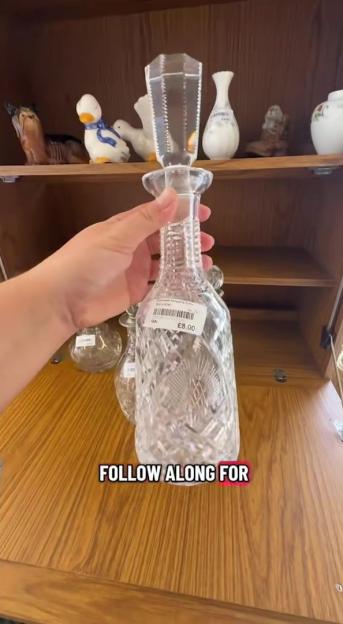 A hand holding a clear glass decanter with an £8.00 price tag, in a charity shop.
