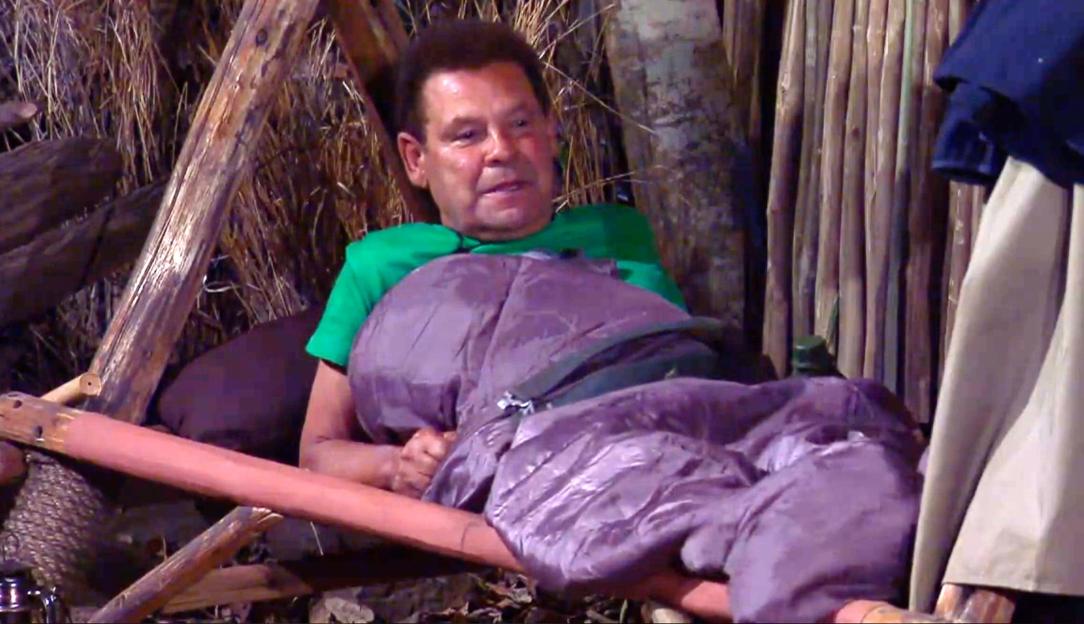 Craig Charles in a green shirt and purple sleeping bag on a wooden cot.