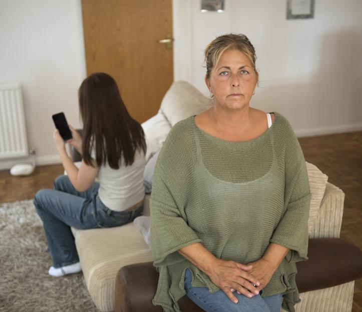 A mother sits facing forward with a concerned expression while her daughter sits behind her on a phone, with her back to the viewer.