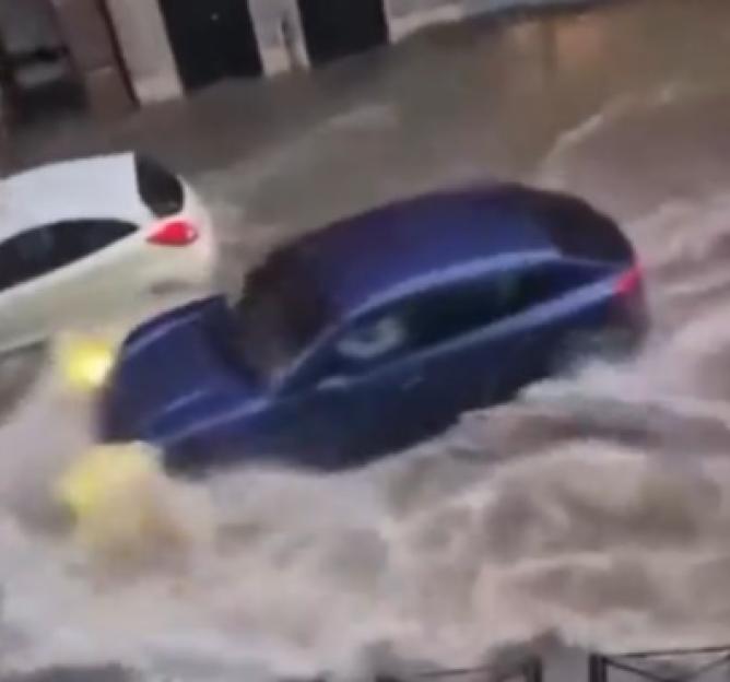 A dark blue car partially submerged in floodwaters next to a white car on a city street.
