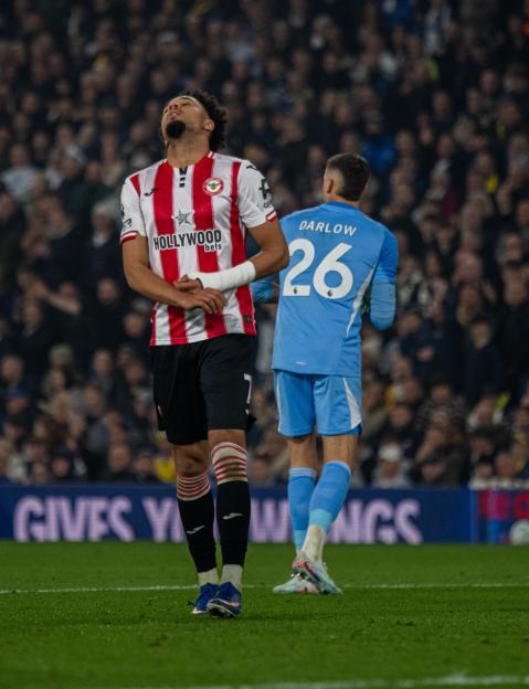 Two soccer players on a field, the one in front in a red and white striped jersey looking up in frustration, the other in a light blue jersey with "DARLOW 26" on the back.