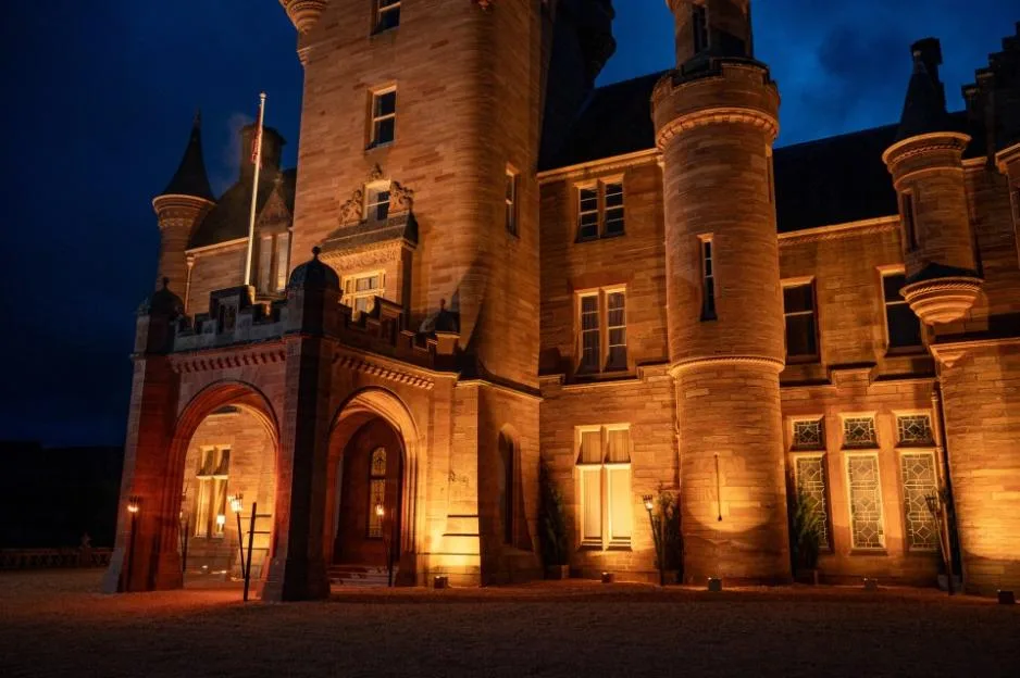 A stone castle with multiple turrets and arches is illuminated by warm lights at night, with the sky a deep blue.