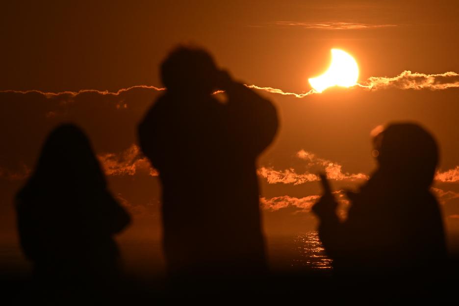 Silhouetted figures observe a partial solar eclipse over the ocean.