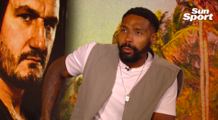 A man with a beard, wearing a white t-shirt and grey vest, speaks into a microphone.