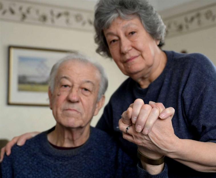 An older woman with gray hair holds the hands of an older man with gray hair, looking at the camera.