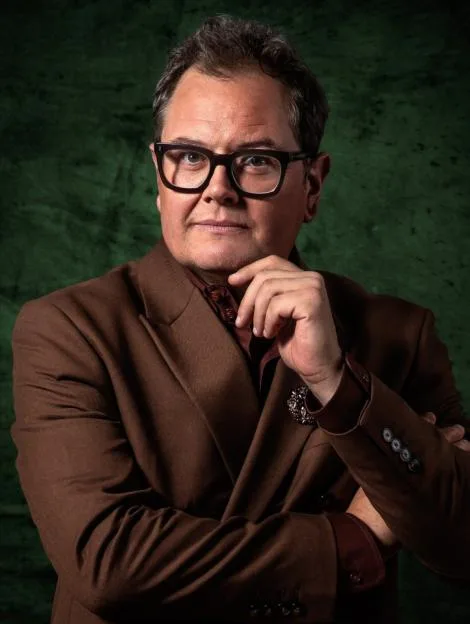 Alan Carr in a gold-framed portrait, wearing a brown suit and glasses, against a dark green background, set on patterned wallpaper above a fireplace mantel with decorative items.