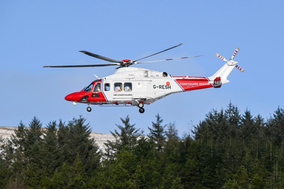 A white and red HM Coastguard helicopter flying over a forest with snow-covered mountains in the background.