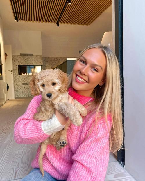 A smiling blonde woman in a pink sweater holds a light brown puppy.