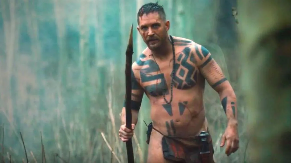 Tom Hardy as James Delaney in Taboo, bare-chested with tribal tattoos, holding a spear in a wooded area.