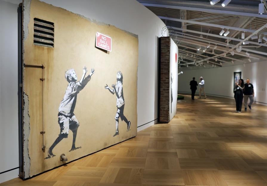 Banksy's "No Ball Games" artwork, depicting two children playing against a wall with a sign saying "NO BALL GAMES," displayed in an art gallery.