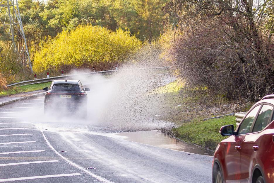A car splashes water from a flooded road onto the side of the road with another car.