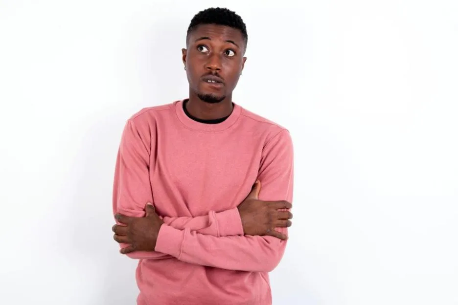 A young man wearing a pink sweater bites his lip and looks worried with crossed arms.