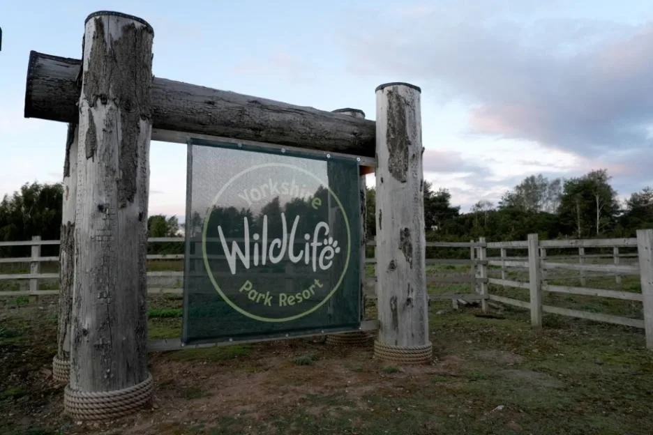 The entrance sign for Yorkshire Wildlife Park Resort, made of thick wooden logs and a hanging mesh banner, set against a backdrop of trees and a cloudy sky.