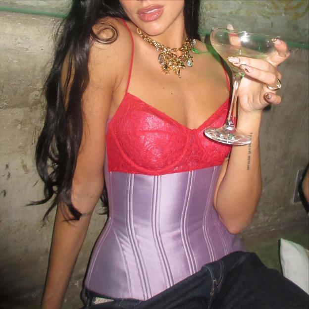 Dua Lipa wearing a red bustier and purple corset while holding a champagne glass.