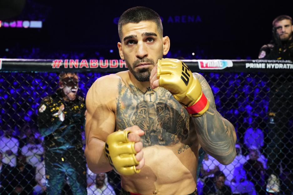 UFC fighter Ilia Topuria in a boxing stance in the ring, wearing gold gloves and displaying chest and arm tattoos.