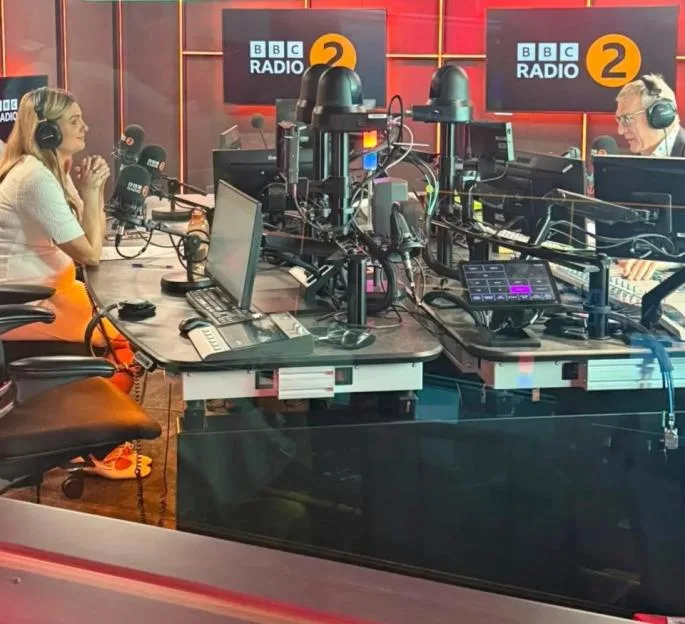 A woman and a man in a BBC radio studio with monitors displaying "BBC RADIO 2".