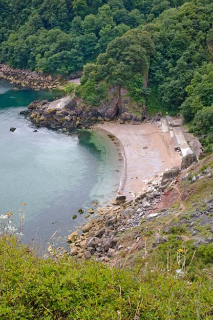 Anstey's Cove in Torquay, South Devon, UK, showing a curved sandy beach with rocks and lush green trees along the coastline.