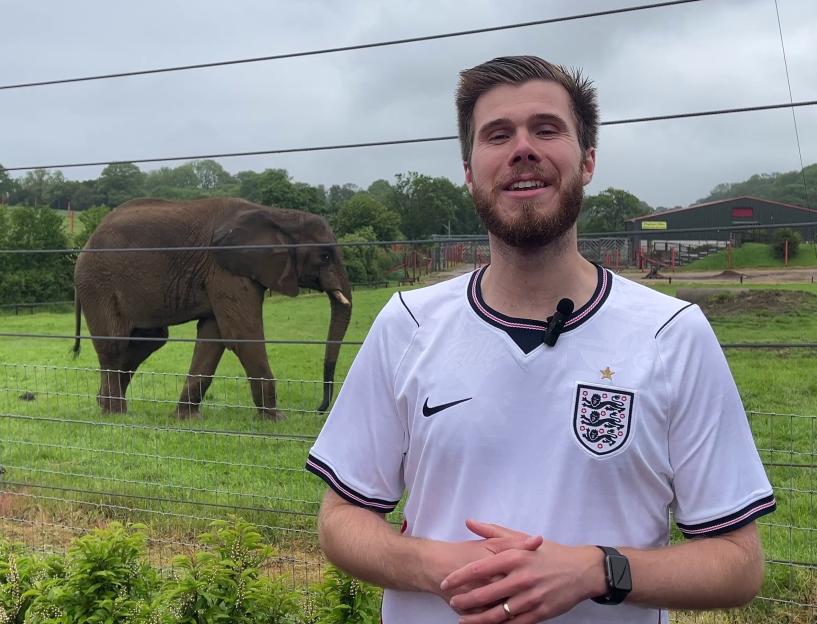 A man in a white England football shirt stands smiling in front of an enclosure with elephants and green hills.
