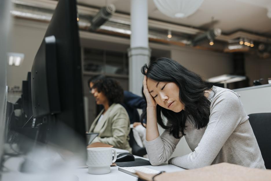 Exhausted female business professional with eyes closed sitting with head in hand near desk at office