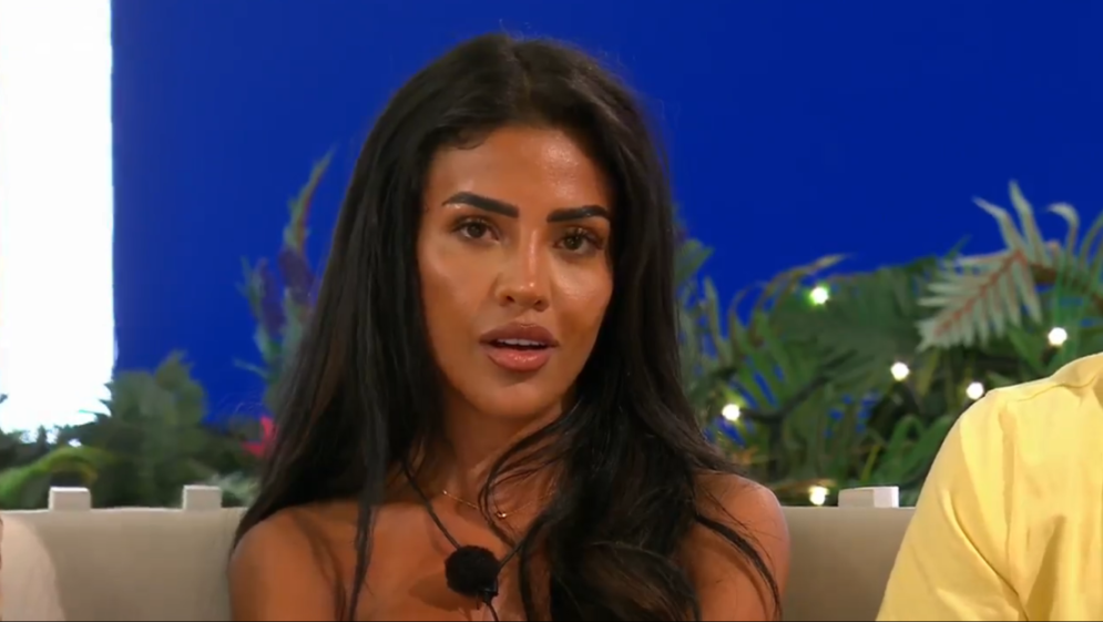 A woman with long dark hair and brown eyes looks slightly to her right, with her mouth slightly open.