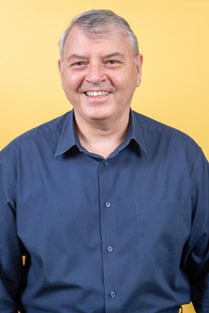 Steve Pharoah, wearing a blue shirt, smiling in front of a yellow background.