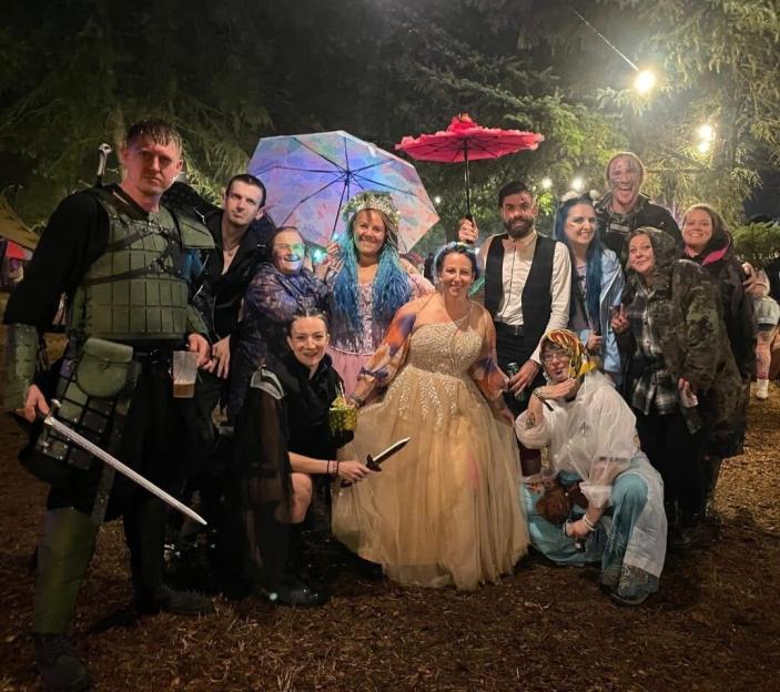 A group of adults in costume, some holding props and umbrellas, posing at an outdoor event at night.